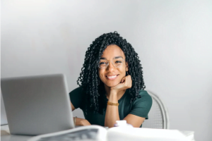 Mulher negra com cabelo cacheado em frente a um notebook e rindo para frente